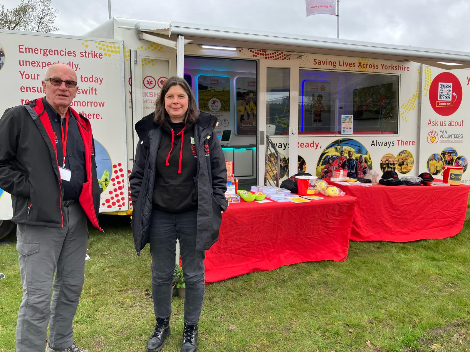 Two people stood on a field in front of a promotional vehicle with two tables that both have red coverings and contain a number of merchandise items.